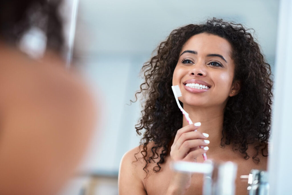 Mulher sorrindo em frente ao espelho enquanto escova os dentes após o uso do clareador dental — cuidados com a higiene bucal para manter os resultados do clareamento e garantir um sorriso saudável e branco.